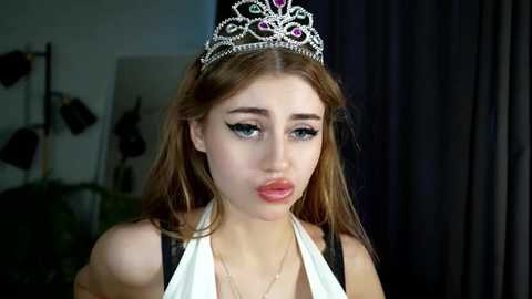 Media: Video of a young woman with fair skin, wearing a silver tiara with pink stones, black eyeliner, and a white halter dress, pouting. Background shows a dark curtain and a blurry studio setup.