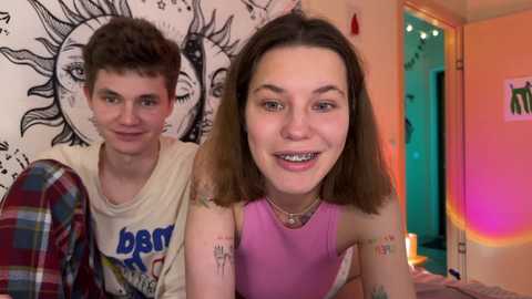 Media: Video of two young people, a smiling girl with braces in a sleeveless pink top, and a boy in a plaid shirt, in a colorful, sun-filled room with sun art and glowing lights.