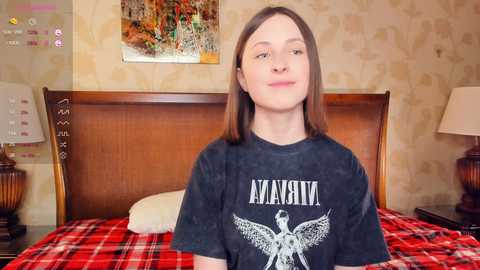 Video of a young woman with straight brown hair, wearing a black Metallica T-shirt, sitting on a red and black plaid bedspread in a bedroom with floral wallpaper and a wooden headboard.