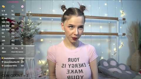 Media: Video of a young woman with fair skin, brown hair in buns, wearing a pink T-shirt with \"YOUR SWEET DREAMS ARE KILLING ME\" printed in black. Background includes a wooden headboard, string lights, and a grey cushion.