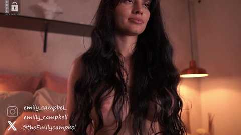 Video of a topless woman with long, dark hair in a dimly lit bedroom. Her skin is light, and she has a serene expression. A lamp and bed are visible in the background.