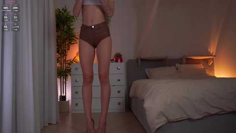 Media: A video of a slender, light-skinned woman in brown shorts and a white top, standing in a dimly lit bedroom with a white dresser, a potted plant, and a bed with beige bedding.