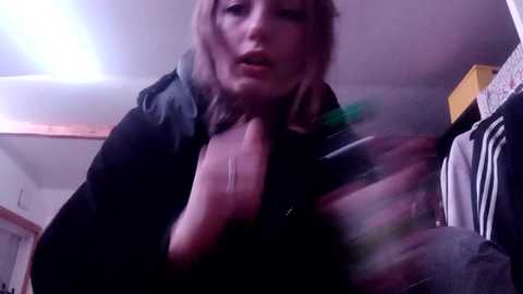 Media: A blurry video of a woman with shoulder-length blonde hair in a black jacket, possibly holding a phone, standing in a dimly lit room with clothes and a yellow box in the background.