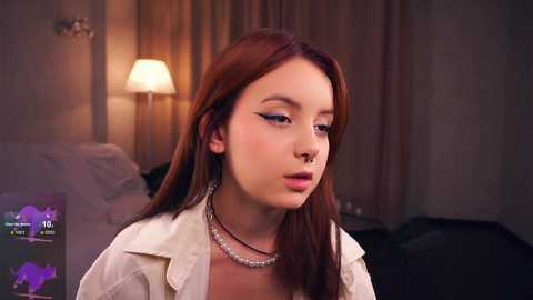 Media: Video of a young Asian woman with long red hair, wearing a white shirt and multiple necklaces, in a dimly lit bedroom with beige curtains and a bedside lamp.