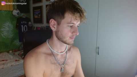 Media: Video of a shirtless young man with light brown hair, wearing multiple necklaces, sitting indoors. Background includes a green inflatable, framed pictures, and a bed.