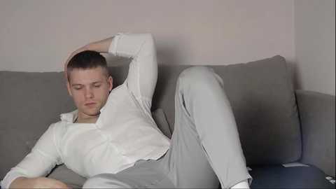 Media: Video of a fair-skinned, short-haired young man with a muscular build, lounging on a grey couch in a casual white button-up shirt and light grey pants, looking relaxed with one arm behind his head.