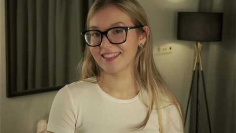 Media: Video of a smiling, light-skinned blonde woman with glasses, wearing a white top, standing in a dimly lit room with a black lamp and curtains.