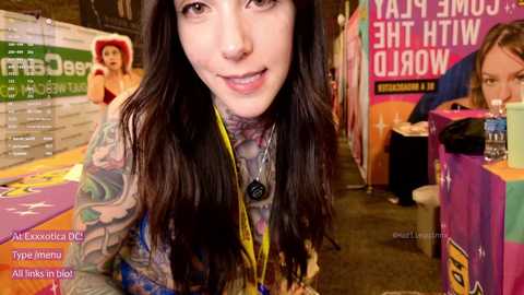 Media: Video of a tattooed woman with long dark hair and a pink lanyard, smiling at the camera in a colorful, crowded convention area.