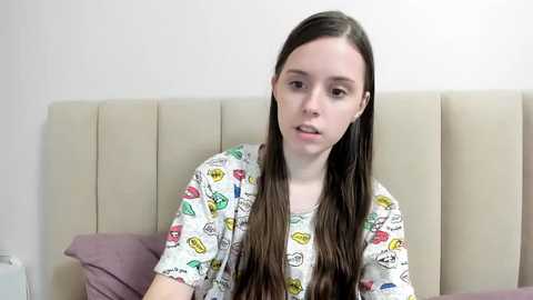 Media: Video of a young Caucasian woman with long brown hair, wearing a cartoon-patterned shirt, sitting on a beige bed with purple pillows, looking slightly off-camera.