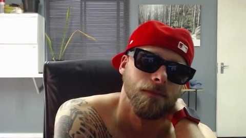 Media: Video of a bearded man in a red baseball cap, black sunglasses, and a black tank top, sitting in a black chair. Background features a white desk, a potted plant, and a monochrome forest video.