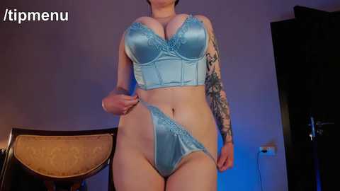 Media: Video of a curvy woman with light skin, large breasts, and tattoos on her arm. She wears a light blue satin bra and matching panties, standing indoors.