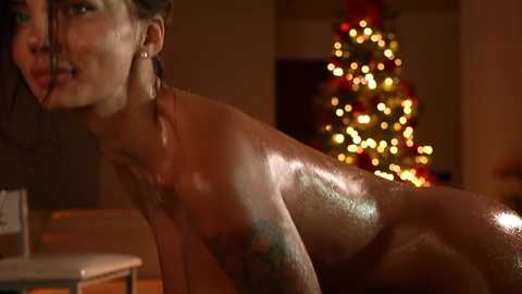 Media: Video of an oiled, nude woman with wet hair, gazing to the left. Background features a dimly lit Christmas tree with golden lights.