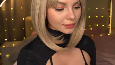 Media: Video of a young woman with straight, shoulder-length blonde hair, wearing a black, high-neck top with cut-out details, against a warm, bokeh background with golden lights.