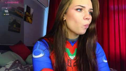 Media: A video of a young woman with long brown hair, wearing a blue and red superhero costume, pouting in a dimly lit room with red curtains.