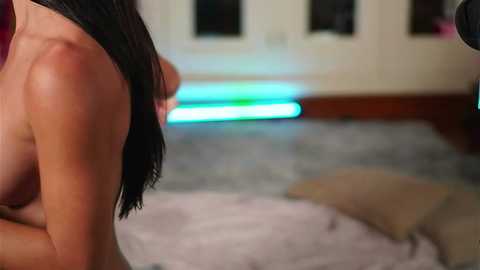 Media: Video of a nude woman with fair skin, medium-sized breasts, and long black hair, kneeling on a gray carpet in a softly lit room with a glowing blue light strip on the floor.