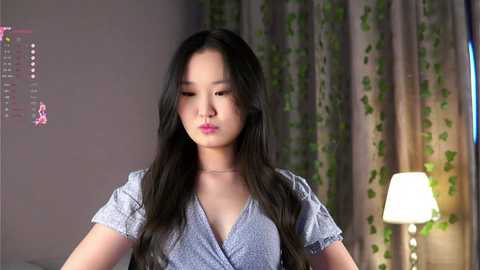 Media: Video of a young East Asian woman with long, dark hair, wearing a light gray, polka-dotted dress, in a softly lit bedroom with a green leaf-patterned curtain and a lamp.