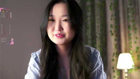 Media: A video of an East Asian woman with long, dark hair, wearing a gray cardigan, smiling softly. She stands in a softly lit room with green curtains and a lamp.