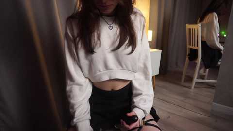 Media: Video of a young woman with long brown hair, wearing a cropped white sweater and black shorts, sitting on a wooden floor in a dimly lit room with a table, chairs, and a lamp in the background.