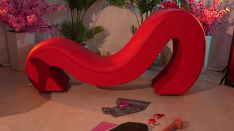 Media: Video of a modern red curved sofa with a sleek design, surrounded by pink blooming flowers in white pots, set against a beige wall, with scattered red and pink items on the floor.