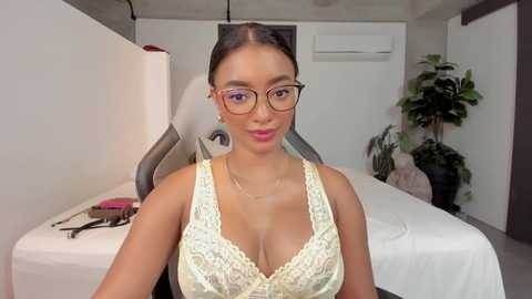 Media: Video of a young woman with light brown skin, wearing glasses and a lace bra, seated on a white bed in a modern, minimalist bedroom with a potted plant and air conditioning unit.