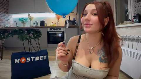 Media: A video of a young woman with long red hair and a tattoo on her right arm, sitting on a wooden floor in a modern kitchen, holding a bottle of beer, wearing a grey strapless top.