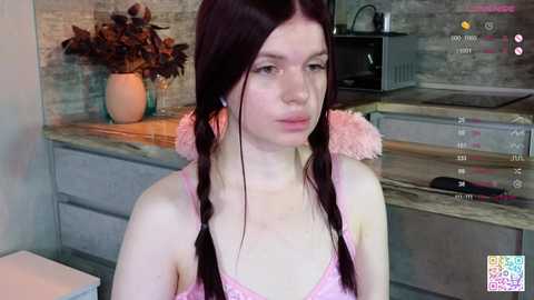 Media: A video of a pale-skinned woman with long dark hair in twin braids, wearing a pink lace bralette, in a modern kitchen with wooden countertops and a beige brick wall.