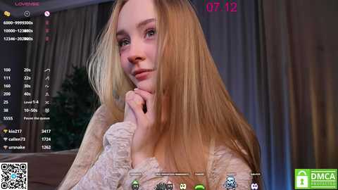 Media: A video of a young Caucasian woman with long blonde hair, wearing a white lace sweater, posing in a cozy room with curtains and a plant. The image is taken from a live stream, showing a chatroom with viewers.