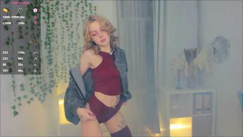 Media: A video of a young, slim blonde woman with shoulder-length hair, wearing a dark red crop top and shorts, standing in a softly lit bedroom with greenery and bookshelves in the background.