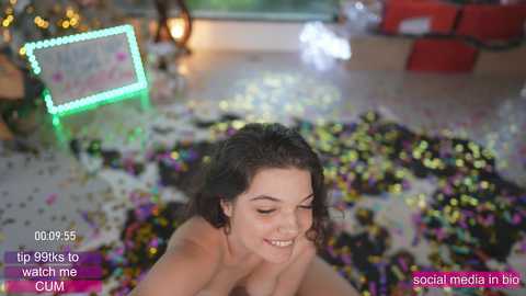 Media: Video of a topless, smiling young woman with curly brown hair, surrounded by confetti, in a festive, dimly lit room with a neon sign and Christmas tree.