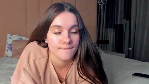 Media: Video of a young woman with long brown hair, wearing a beige top, smiling softly, in a cozy bedroom with a bed and pink curtains in the background.