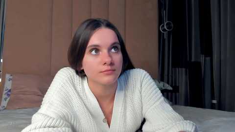 Media: Video of a young Caucasian woman with straight brown hair, wearing a white knitted cardigan, lying on a bed with beige headboard, looking contemplative.