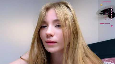 Media: Video of a young Caucasian woman with long, straight blonde hair and fair skin, wearing a light pink top, smiling slightly, in a room with a white wall and a pink and white patterned bedspread.