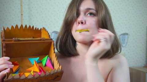 Media: Video of a young, topless woman with shoulder-length brown hair, holding a wooden box with colorful origami inside, yellow tape over her mouth, in a simple room with wallpaper and light switches.