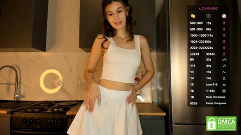 Media: Video of a young Asian woman with shoulder-length brown hair, wearing a white crop top and skirt, standing in a modern kitchen with stainless steel appliances and a marble backsplash.