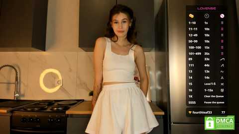 Media: Video of a young woman with long brown hair, wearing a white crop top and skirt, standing in a modern kitchen with stainless steel appliances, a marble backsplash, and a glowing circular light fixture.