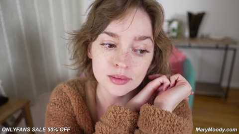 Media: Video of a young woman with light skin, freckles, and shoulder-length brown hair, wearing a brown knit sweater, resting her chin on her hand, looking introspective. Background includes a white wall, a window, and a wooden table.