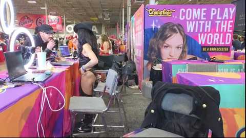 Media: Video of a colorful, crowded indoor event with a large \"Come Play with the World\" banner, featuring a woman in a black dress and hat, and a man in a hat. Tables, chairs, and a ring light are visible.