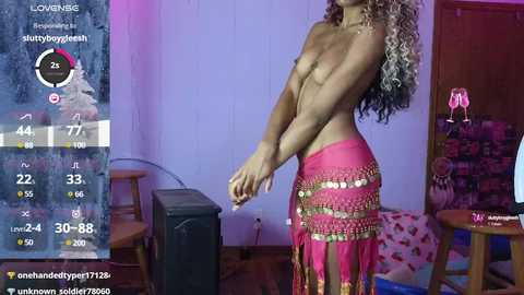 Media: Video of a topless, slender, light-skinned woman with curly, multicolored hair, wearing a pink, beaded skirt, standing in a dimly lit, cluttered room.