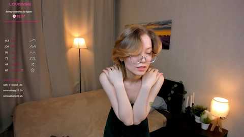 Media: Video of a young woman with glasses and shoulder-length blonde hair, wearing a black dress, standing in a dimly lit bedroom with beige walls and a lit lamp on a nightstand.