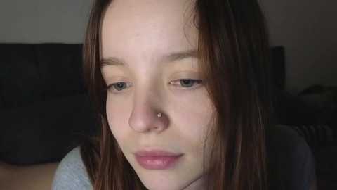 Media: A close-up video of a young woman with light skin, straight brown hair, and a small nose stud. She has a neutral expression, wearing a gray top, set against a dark background.