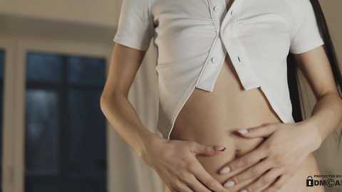 Media: Video of a slender, fair-skinned woman with a small bust, wearing an unbuttoned white blouse, partially exposing her flat stomach and smooth skin, standing indoors by a window.