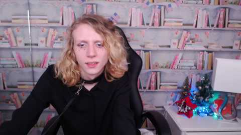 Media: Video of a young woman with shoulder-length blonde hair, wearing a black top, seated in an office chair in front of a bookshelf filled with colorful books.