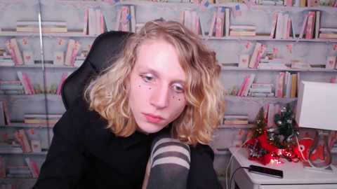 Media: Video of a young woman with curly blonde hair, wearing a black hoodie and striped socks, sitting in a chair against a bookshelf backdrop.