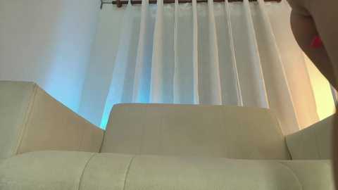 Media: Video of a beige leather sofa with a white curtain in the background. The curtain has vertical stripes of light blue and white. The image is taken from a low angle, emphasizing the sofa's soft texture.