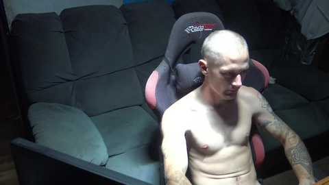 Media: Video of a bald, tattooed, shirtless man sitting on a dark gray gaming chair, in a dimly lit room with a dark sofa and wooden floor.