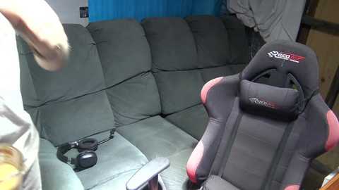 Media: Video of a living room with a gray sectional sofa, a black gaming chair, and headphones on the floor. The background features a blue curtain and a wooden door.