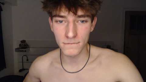 Media: Video of a young, fair-skinned, shirtless male with short brown hair, wearing a thin chain necklace, in a dimly lit, cluttered room with white walls, a small desk, and a door in the background.