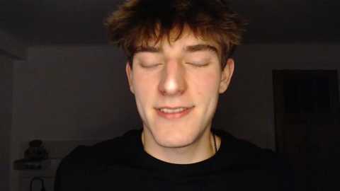 Media: A young man with short, tousled brown hair and fair skin, wearing a black shirt, appears to be closing his eyes and smiling. The background is dimly lit, with a plain, white wall and a door to the right.