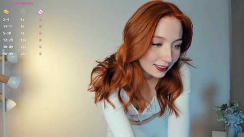 Media: Video of a smiling, fair-skinned woman with long, wavy red hair, wearing a white top, standing in a softly lit room with a modern lamp and gray wall.