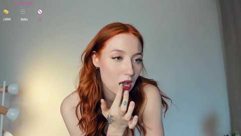 Media: Video of a fair-skinned woman with long, wavy red hair, nude, licking her fingers, in a softly lit room.